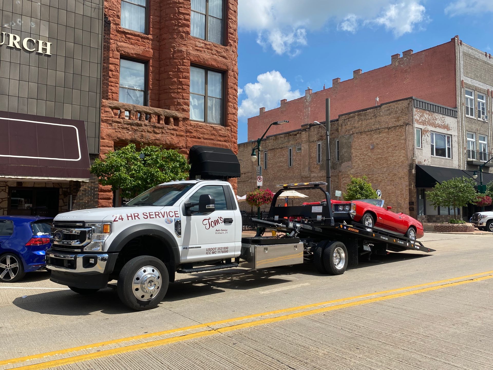 Tow truck with a red car on the flatbed, parked on a street in front of brick buildings.