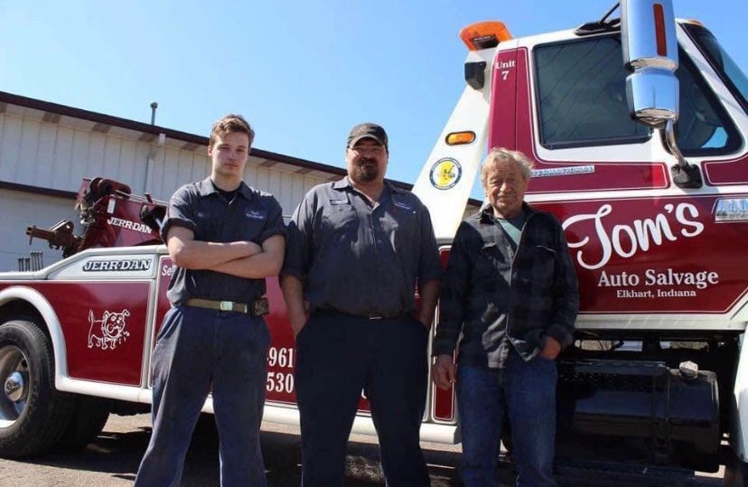 Three men posing in front of a tow truck; the truck is red and white, labeled 