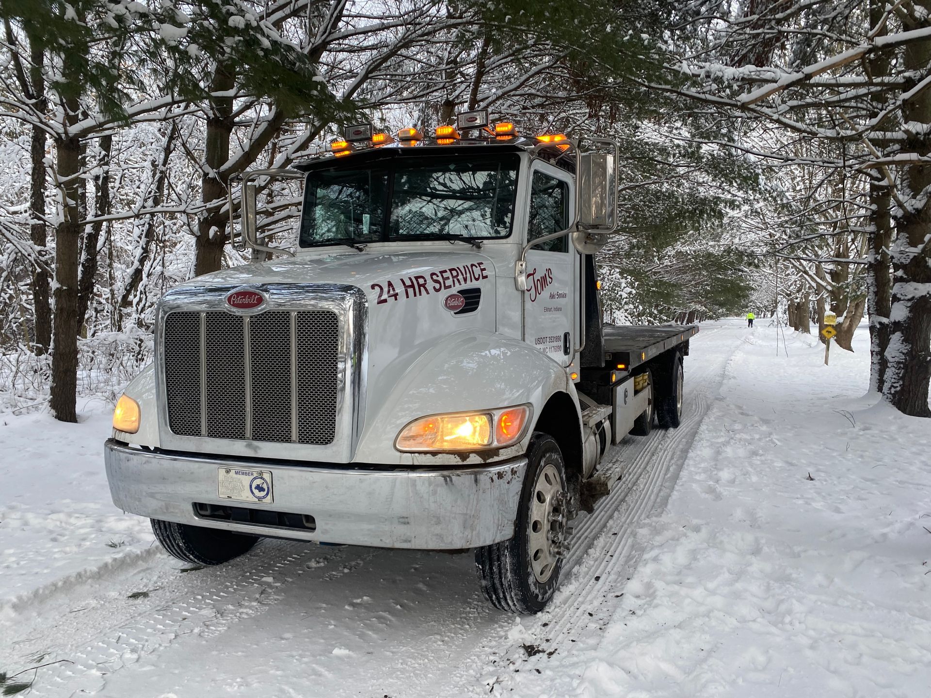 White tow truck on snow-covered road, surrounded by trees.