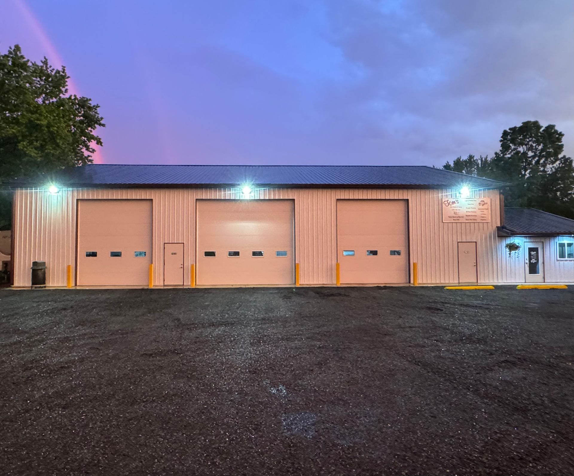 A commercial garage building with three bays, overhead doors, and lights, under a twilight sky with a rainbow.