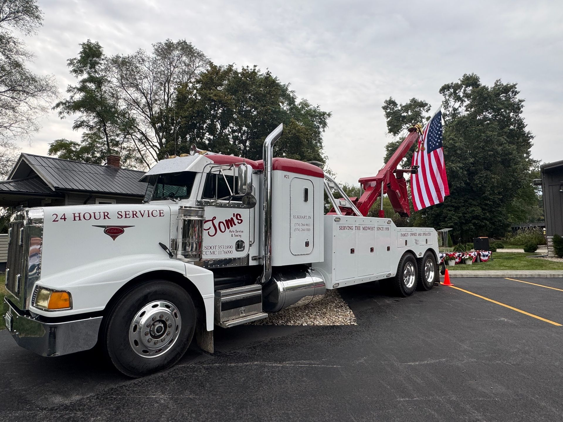 White tow truck with American flag, parked next to a building and trees. 