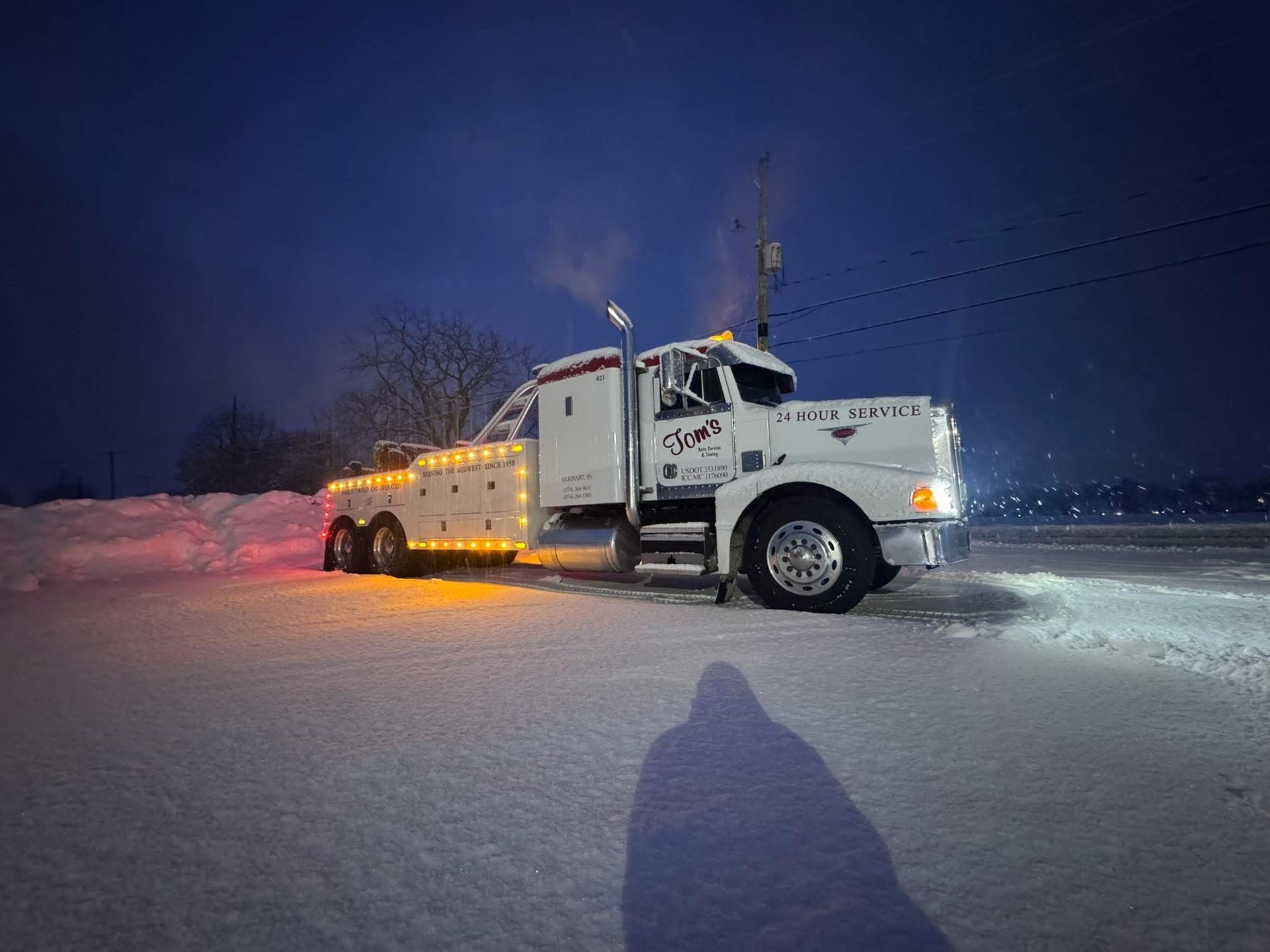 White tow truck in snowy field at night, illuminated by lights, with steam.