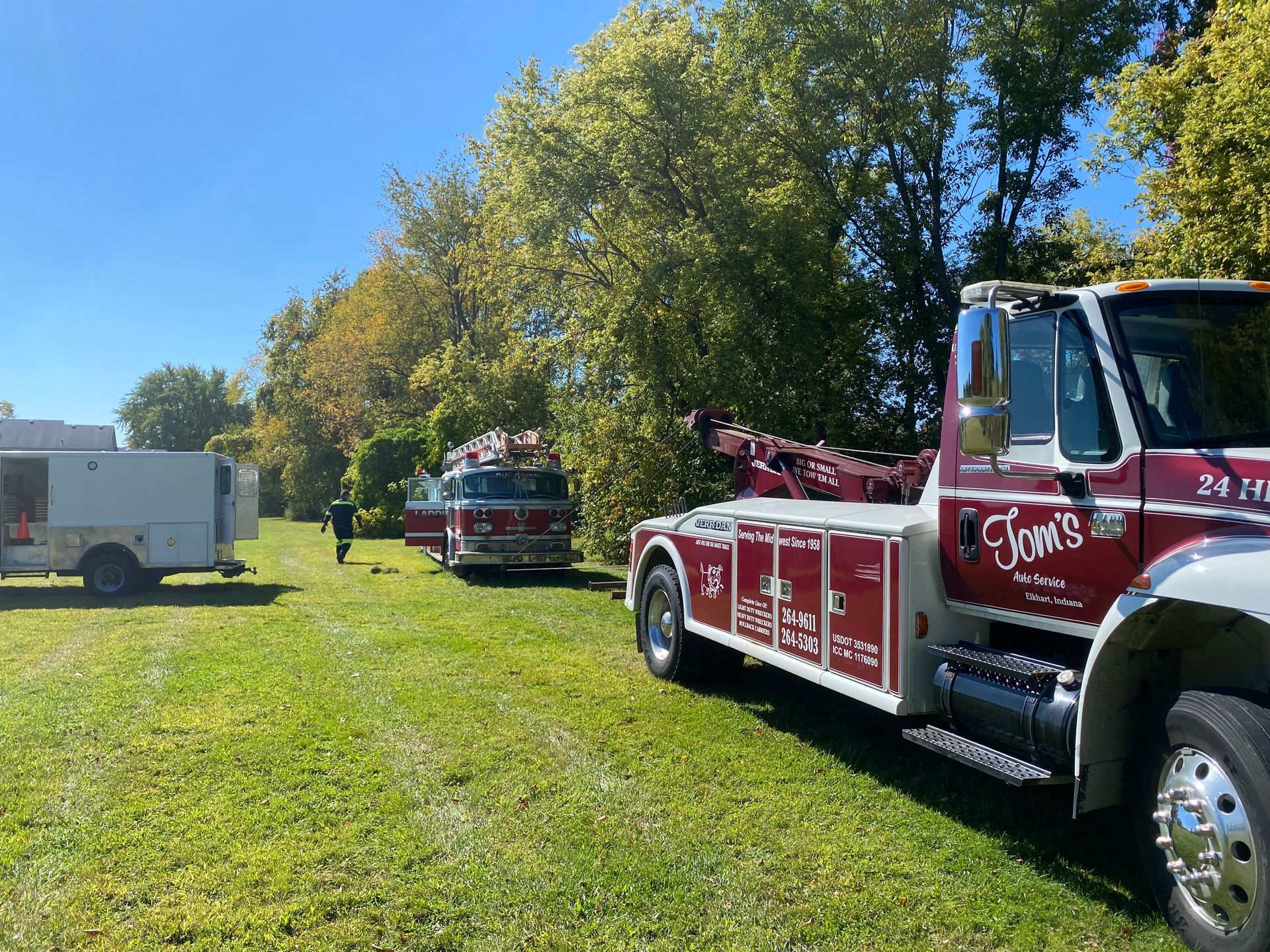 Tow truck, fire truck, and trailer parked on a grassy field, with trees in the background under a blue sky.