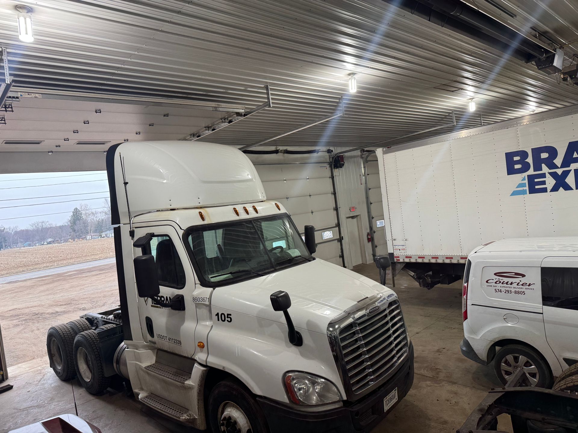 White semi-truck parked inside a garage. A trailer is visible behind it, along with a white van.