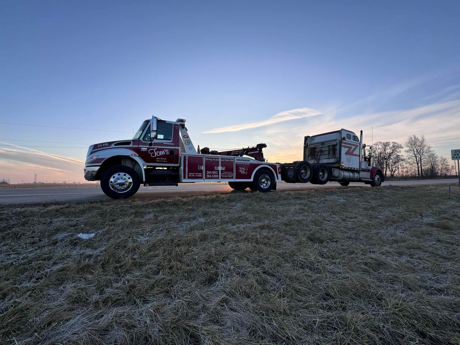 Red tow truck hauling a semi-truck on a grassy roadside at dawn.