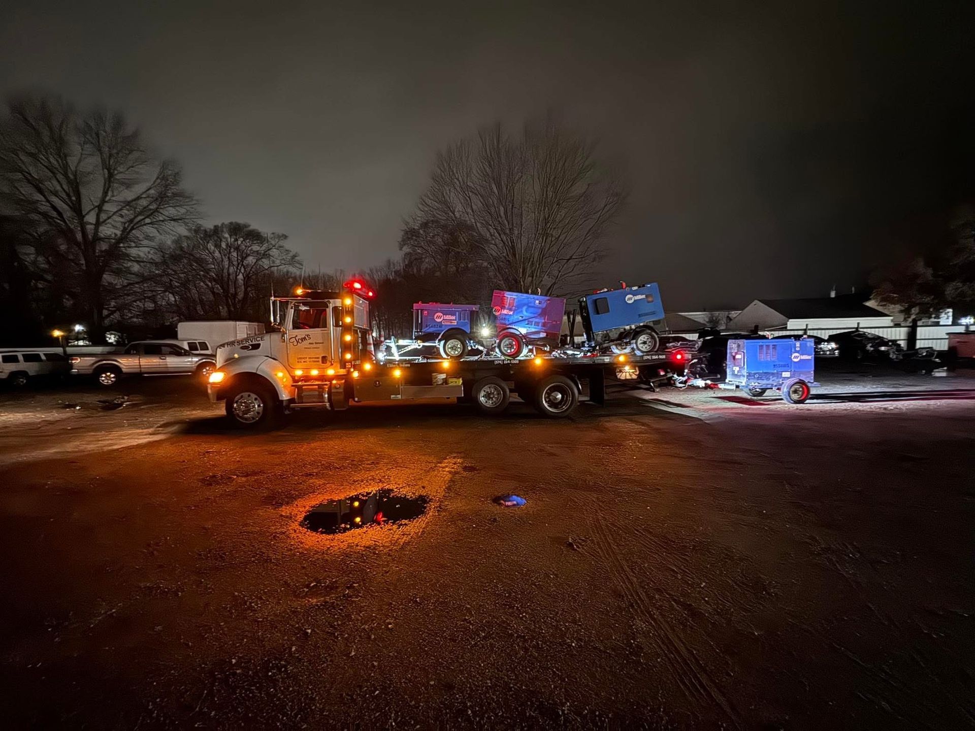 A semi-truck with equipment on its flatbed at night, illuminated by lights in a snowy environment.