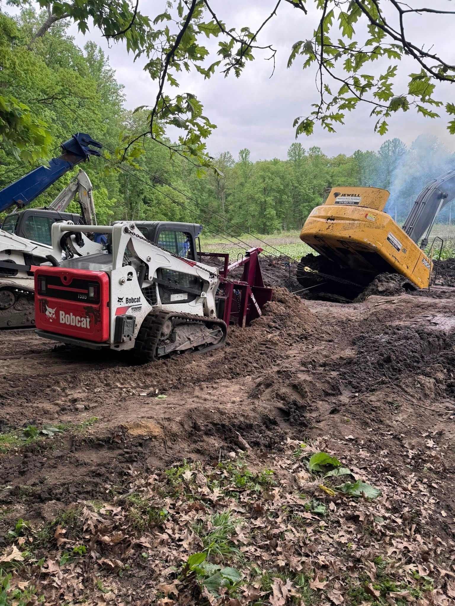 Two skid steers working in muddy terrain; trees in background; overcast sky.