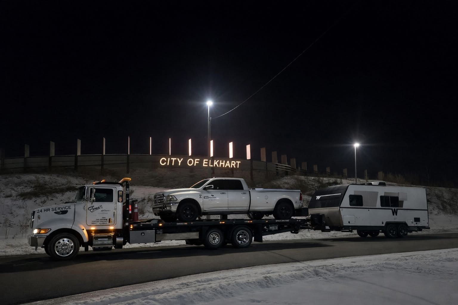 Tow truck hauling a pickup truck and small camper at night in front of the 