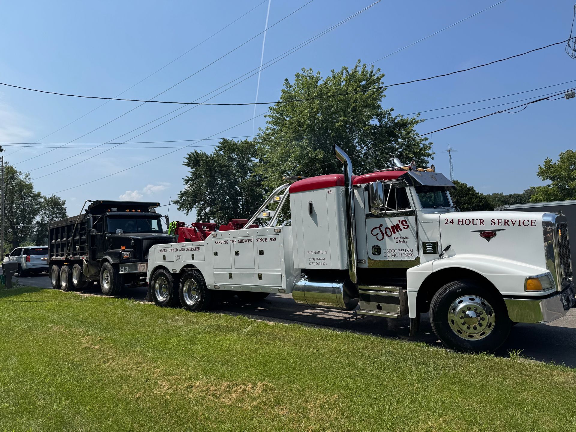 White tow truck pulling a black dump truck on a grassy area, under a blue sky.