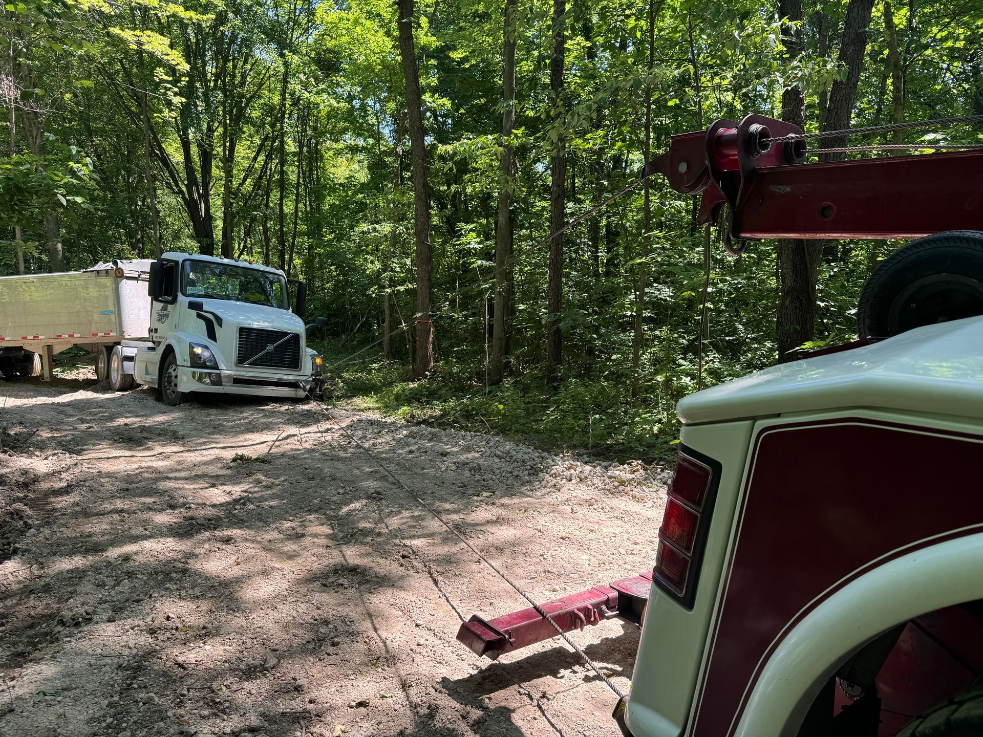 Tow truck towing a semi-truck out of a wooded area on a gravel road, sunny day.
