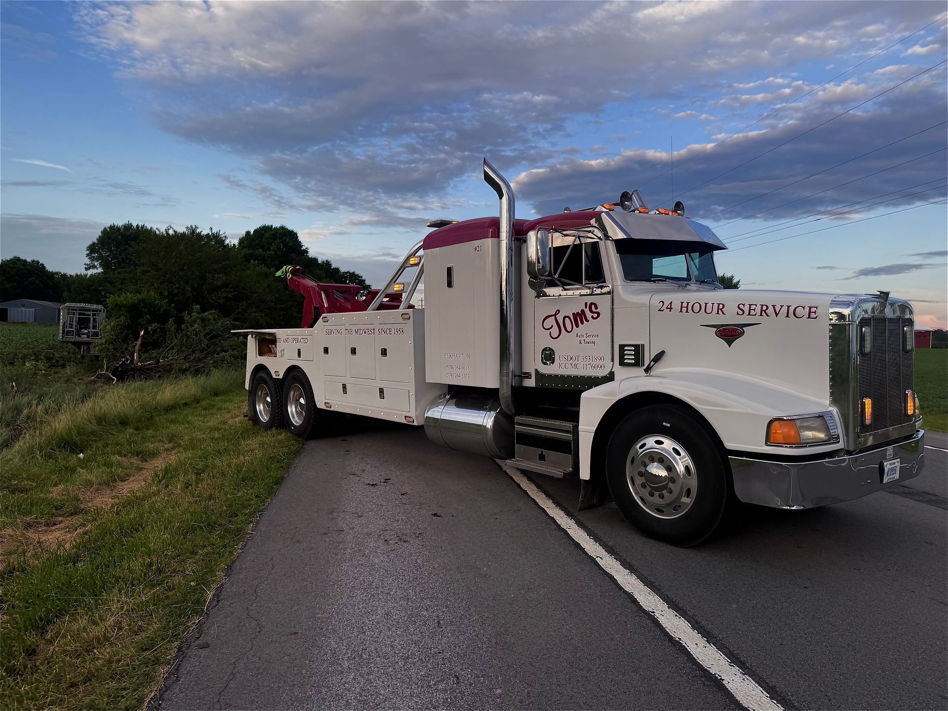 Tow truck on a road, white with maroon accents. Cloudy sky, grass and trees in background.