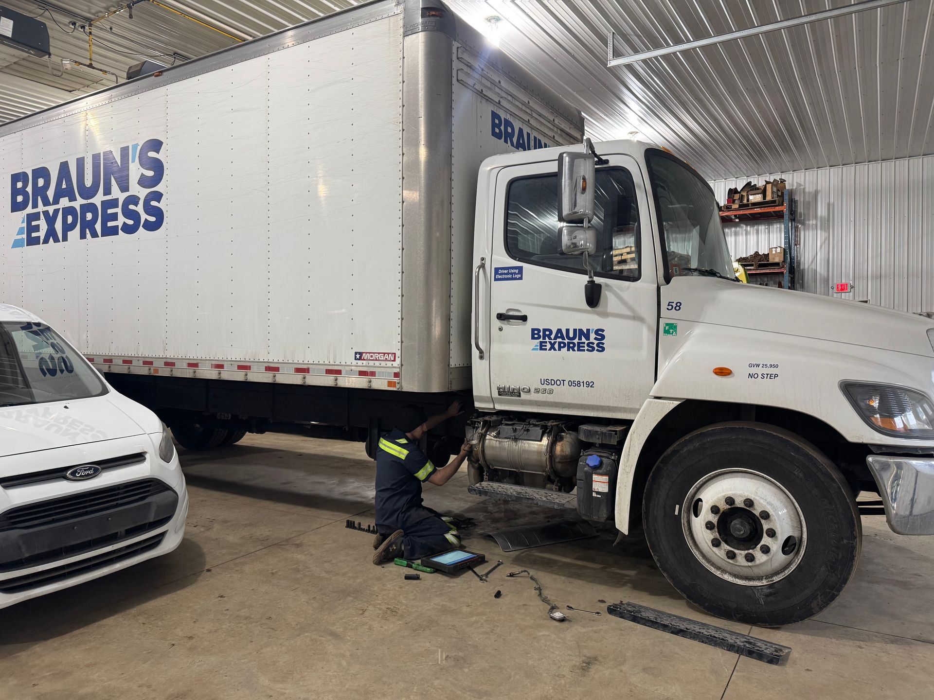 A mechanic works on a white Braun's Express truck in a garage; a white car is parked nearby.