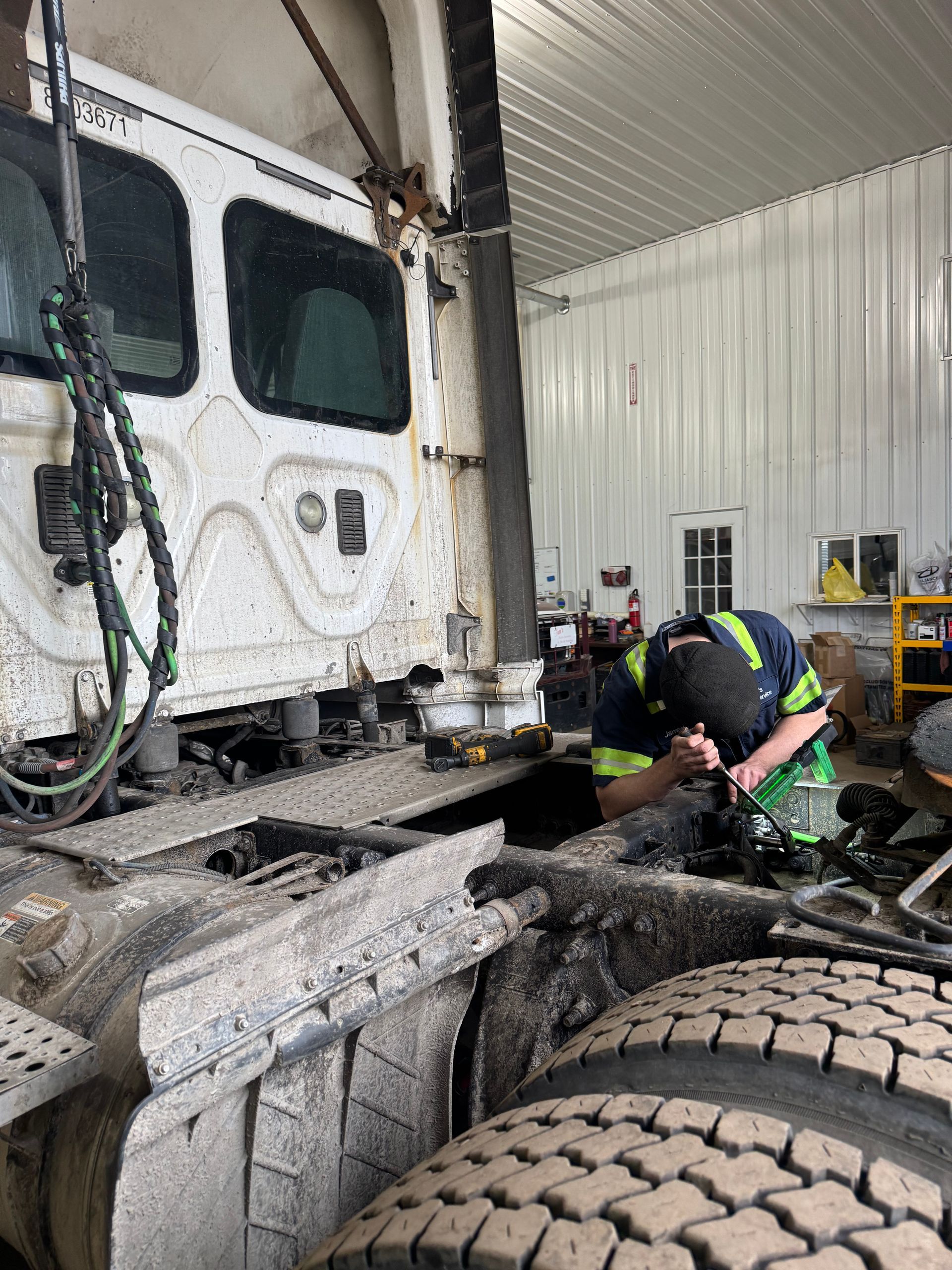 Mechanic works on truck in a garage. Truck cab is white, covered in dirt. The mechanic wears a green and yellow shirt.
