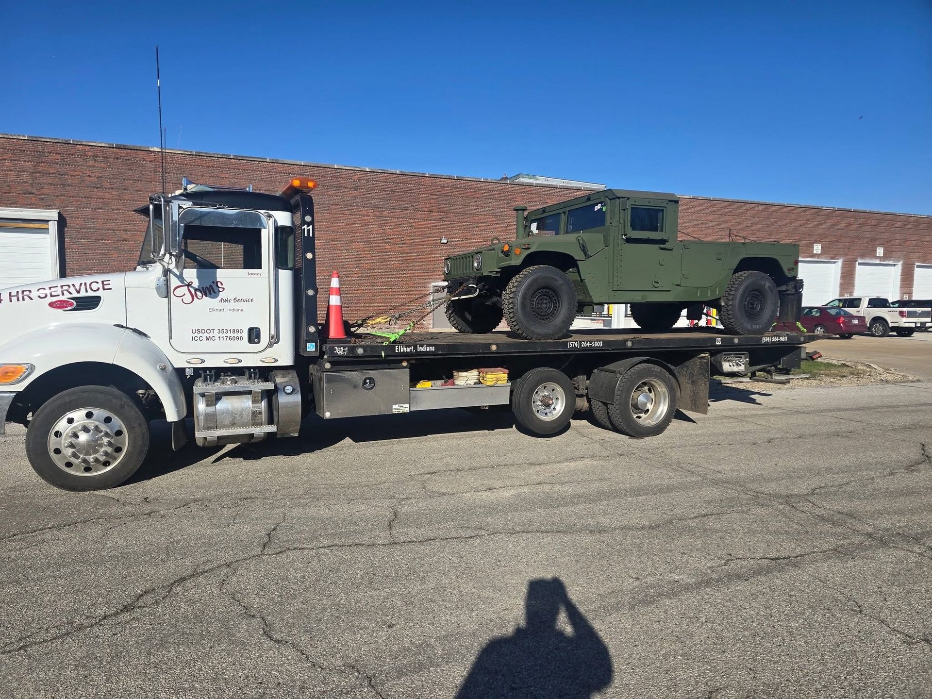 A white flatbed tow truck transporting an olive-green military Humvee parked on an asphalt lot against a brick building.