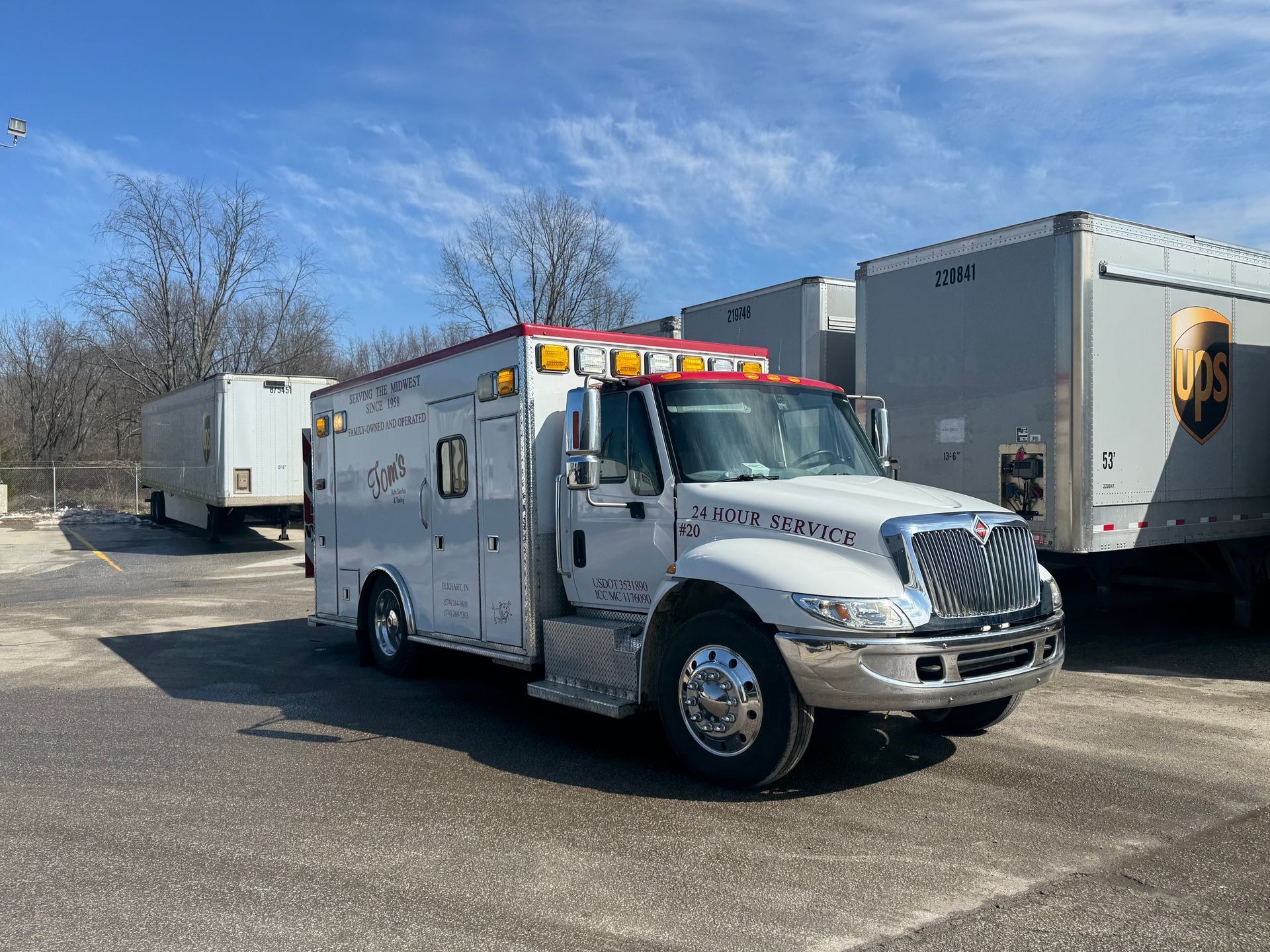 A white ambulance parked in a lot, with two large shipping trailers nearby under a clear blue sky.
