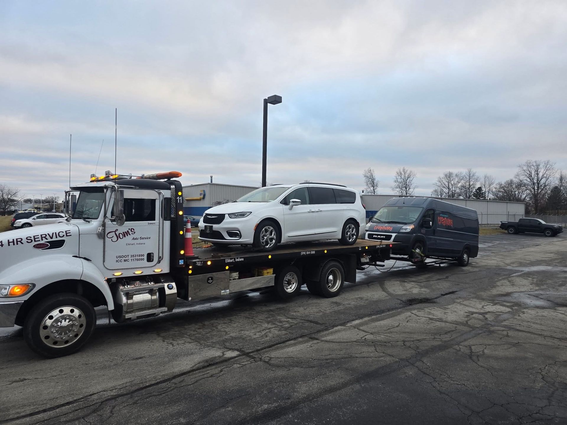 Tow truck carrying a white car and towing a black van, parked outdoors under a cloudy sky.
