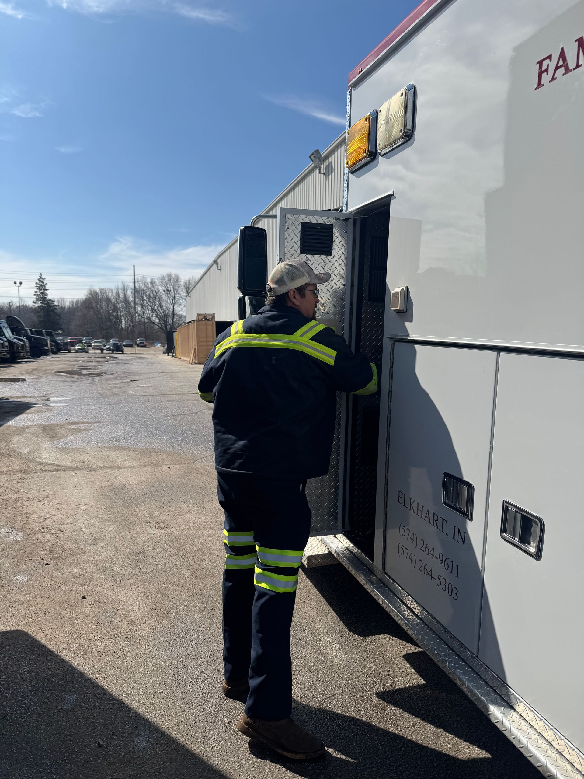 A person in a high-visibility uniform reaching into the back of a large white vehicle parked on a gravel lot.