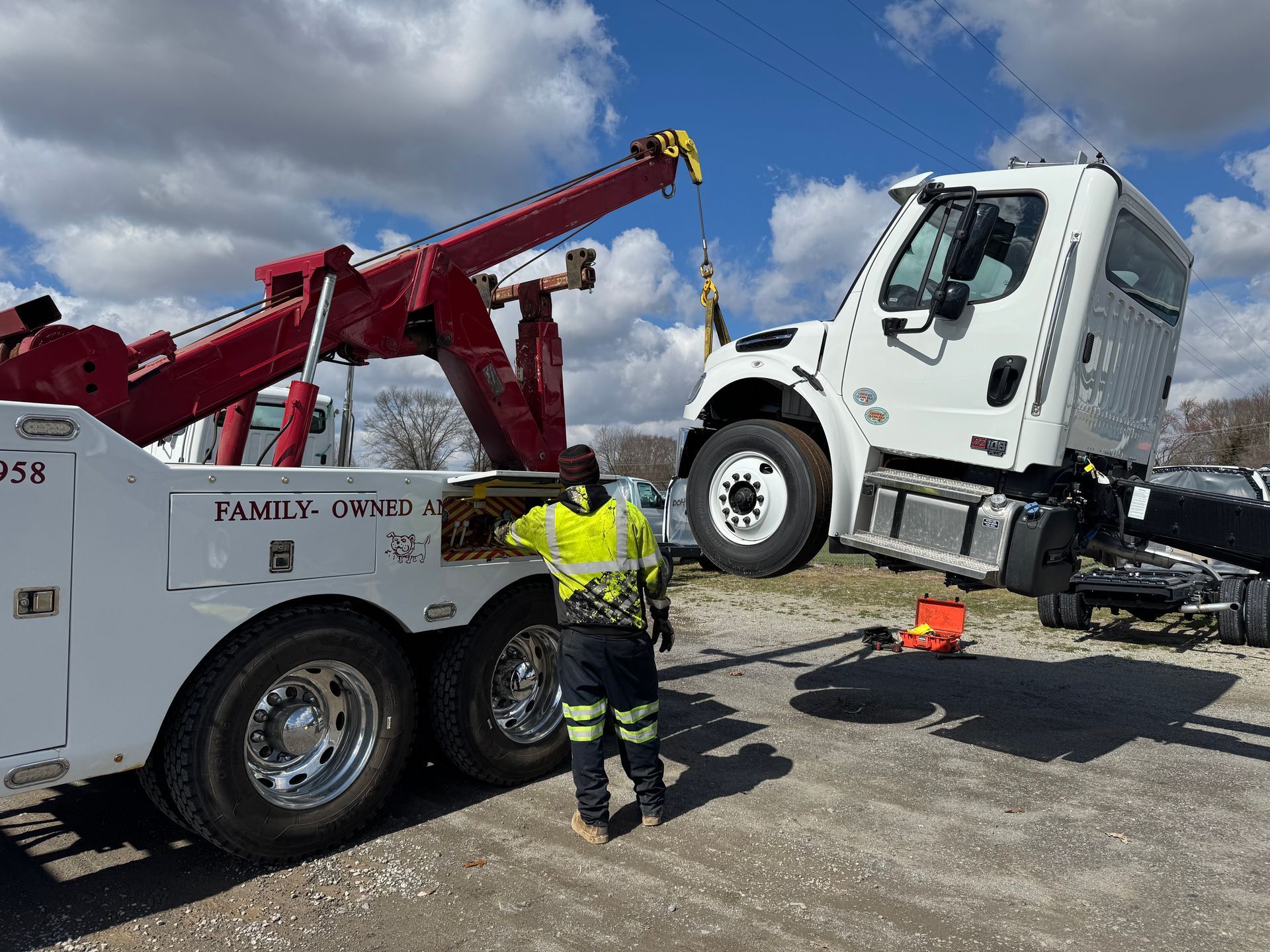 A tow truck operator in a high-visibility jacket uses a red boom crane to lift the cab of a white semi-truck.