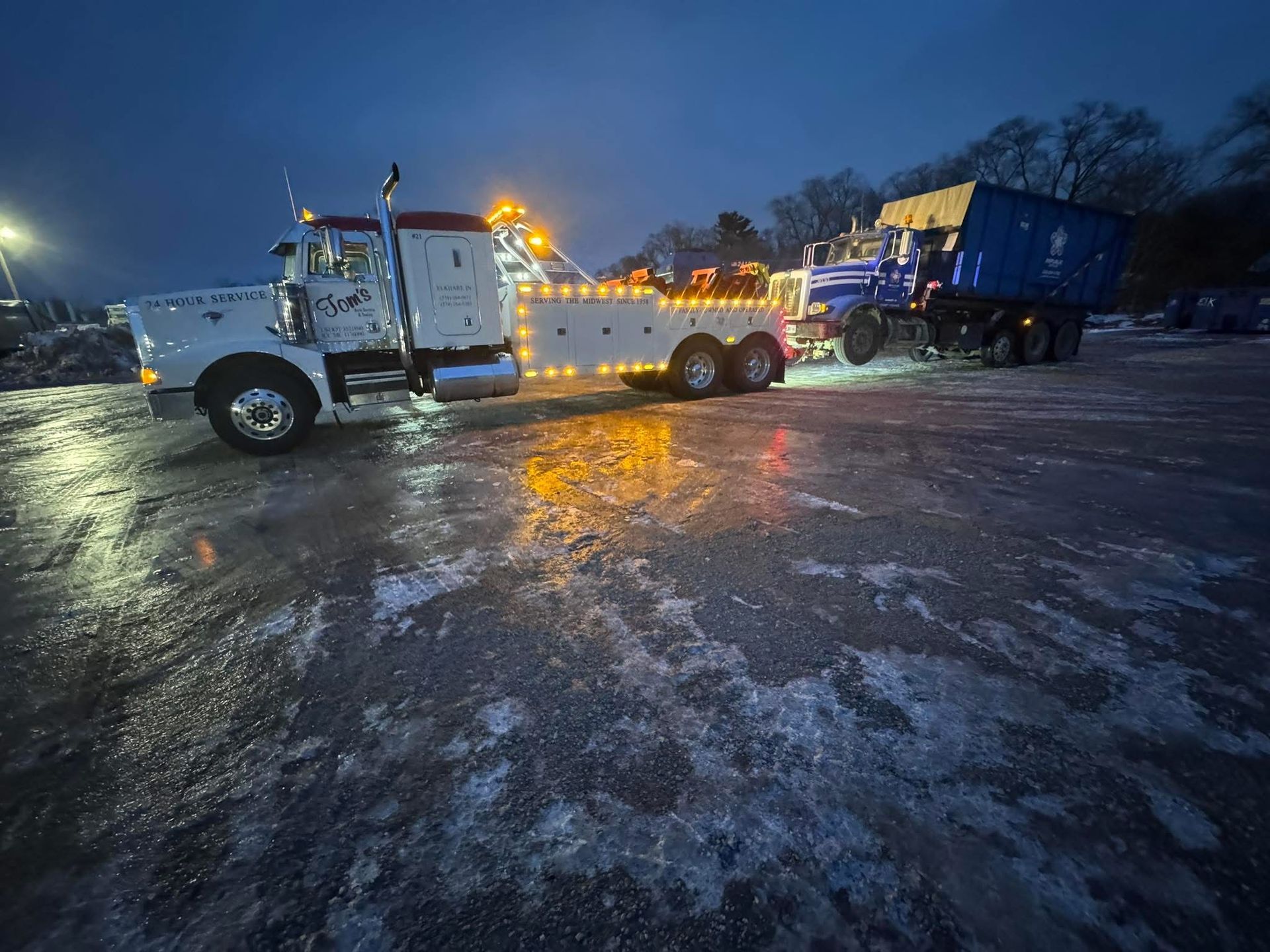 White tow truck towing a blue container in a wet parking lot at night.