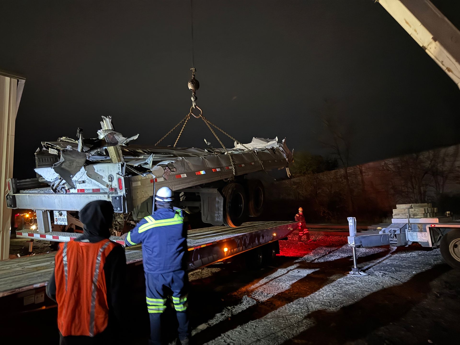 A damaged truck being loaded onto a flatbed at night. Workers in safety gear oversee the crane lifting the wreckage.