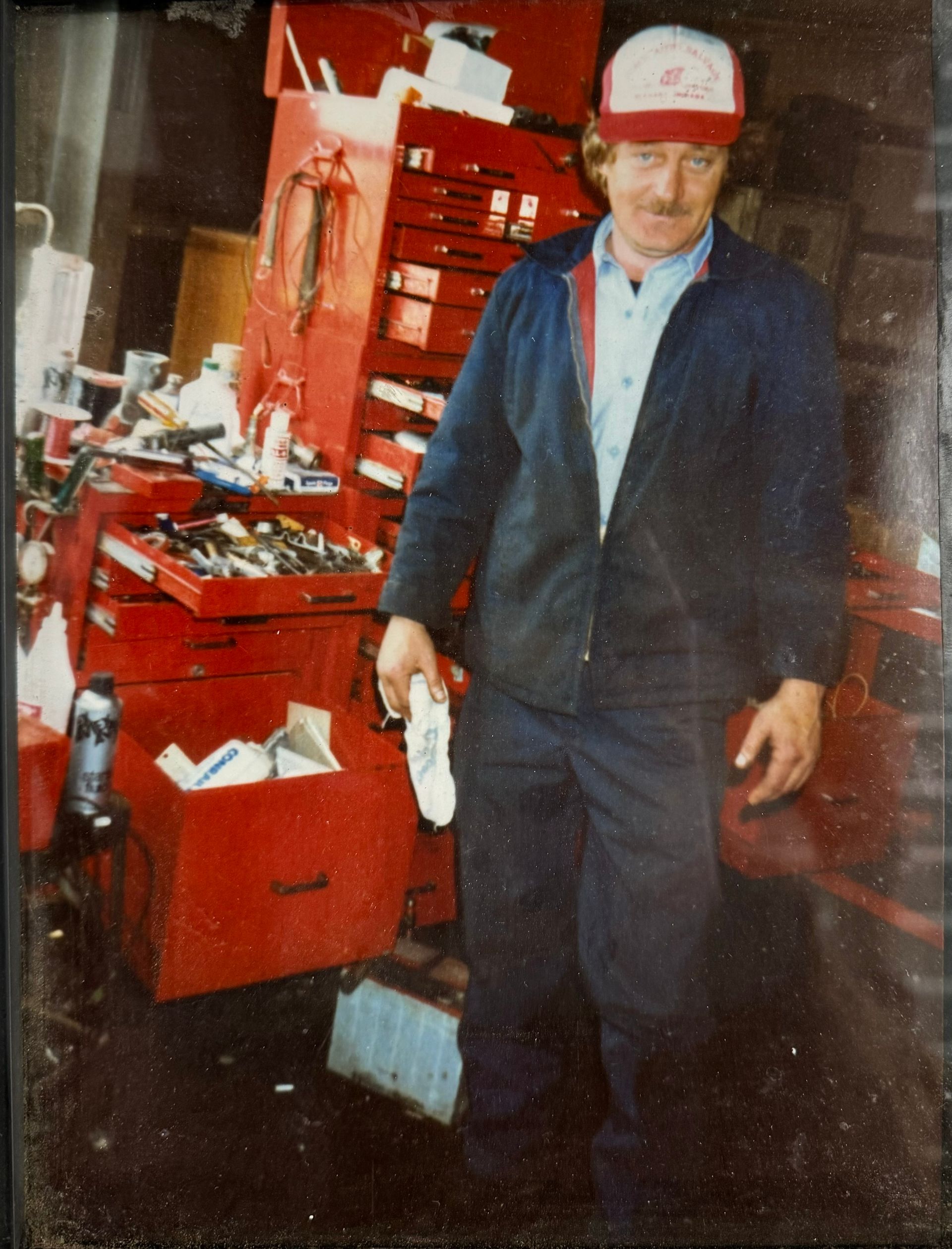 Mechanic in a shop, wearing a cap and jumpsuit, standing near a red toolbox.