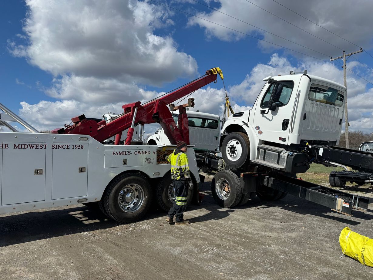 A tow truck operator in high-visibility gear prepares to lift the front of a white truck chassis on a gravel lot.