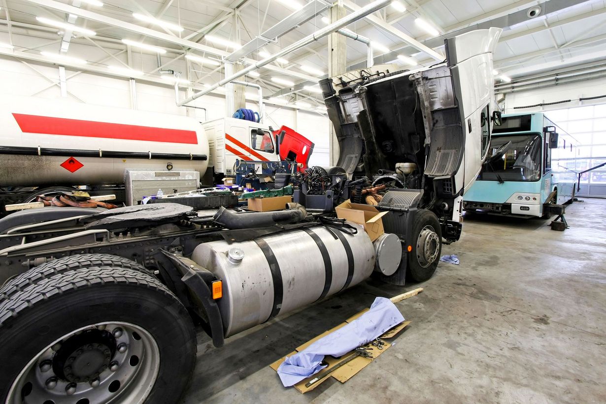 Truck with open hood being serviced in a garage, next to a fuel tank and a bus.