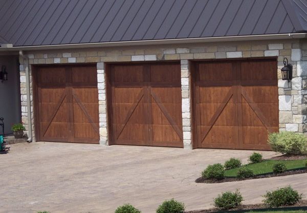 Three wooden garage doors on a stone-and-brick house with a paved driveway and small shrubs