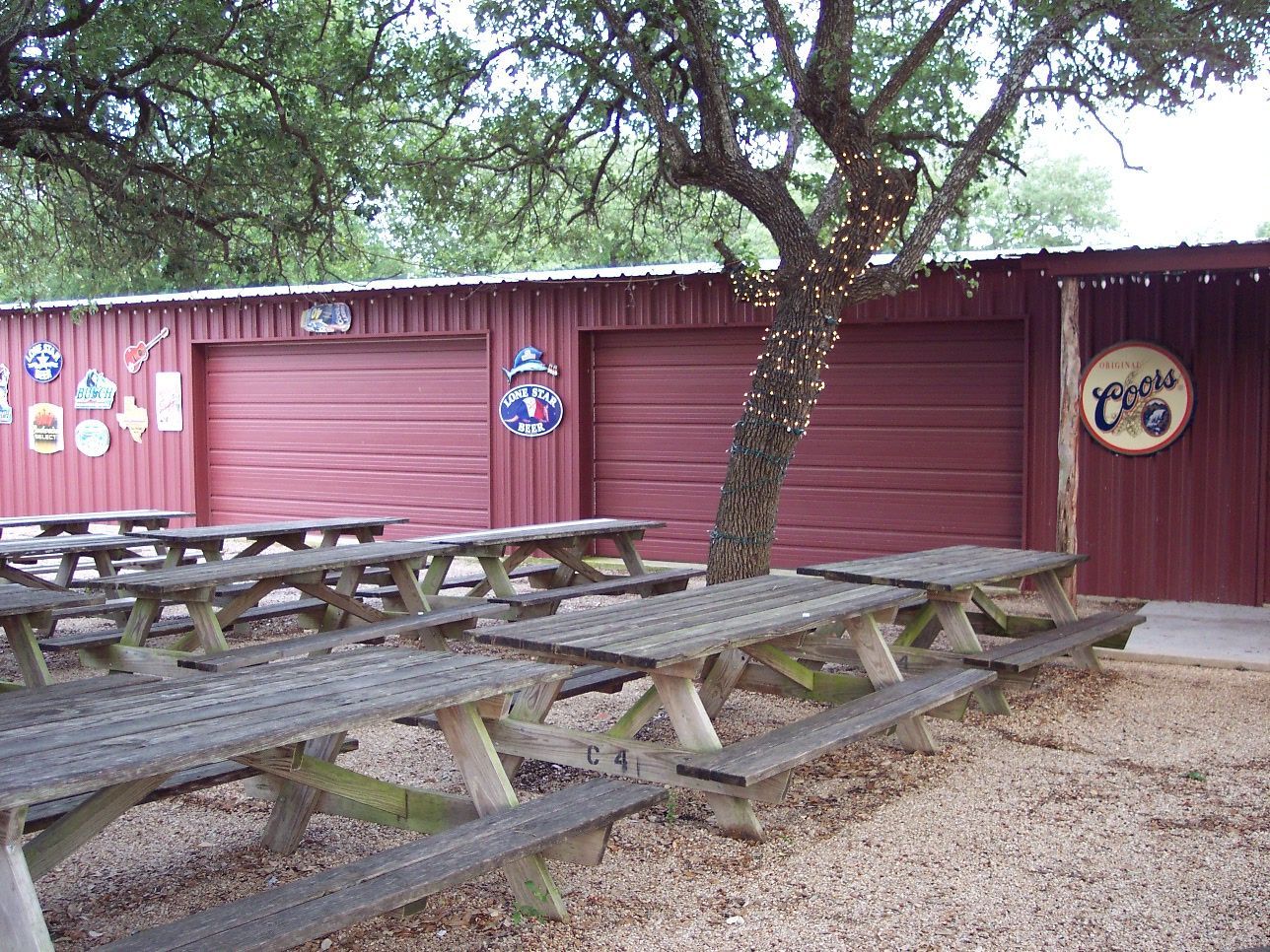 Picnic tables under trees beside a red wooden building with a door and wall decorations