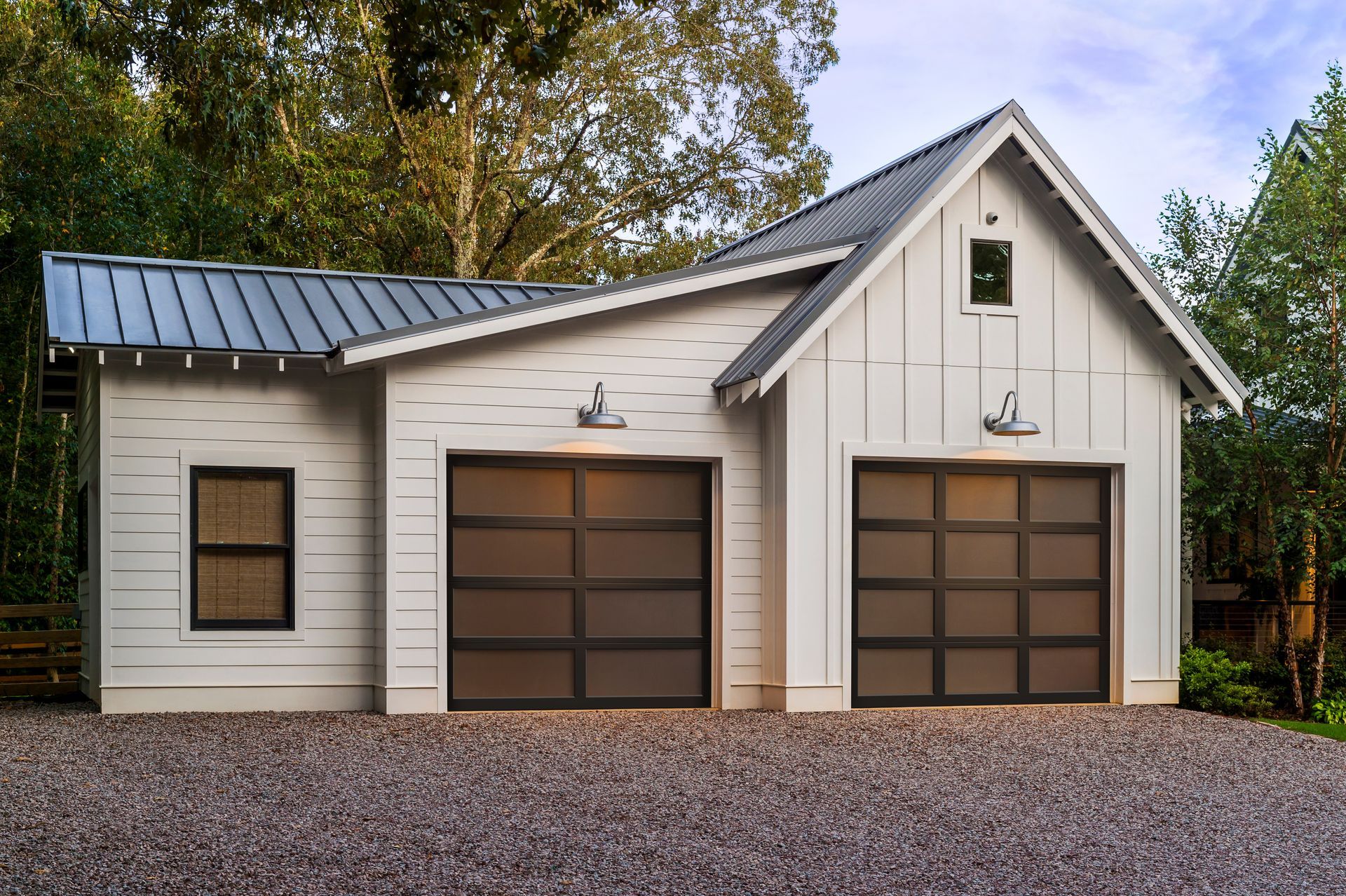 White two-car garage with metal roof and front door, surrounded by trees and gravel driveway