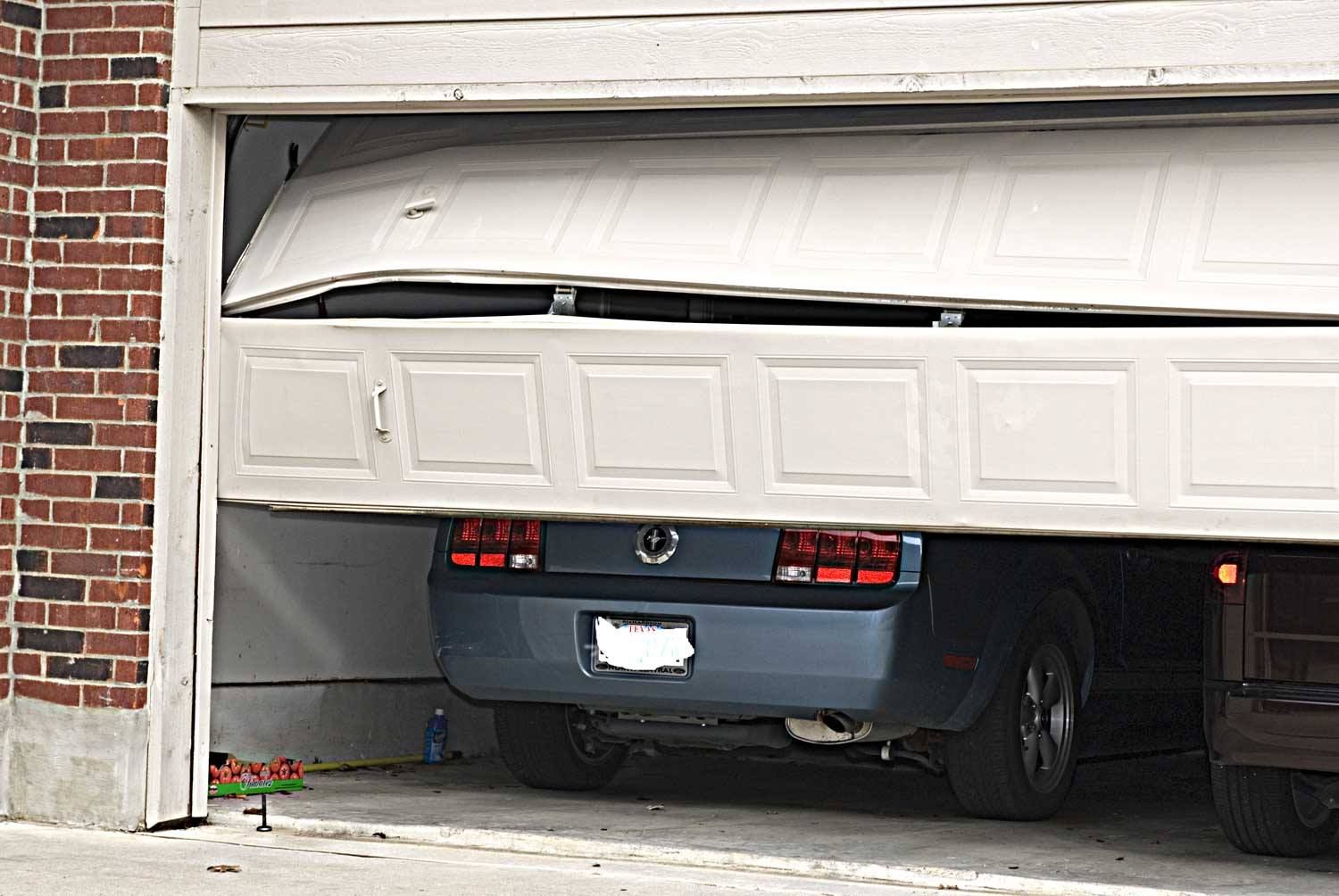 Blue car trapped under a partially closed garage door in a brick driveway