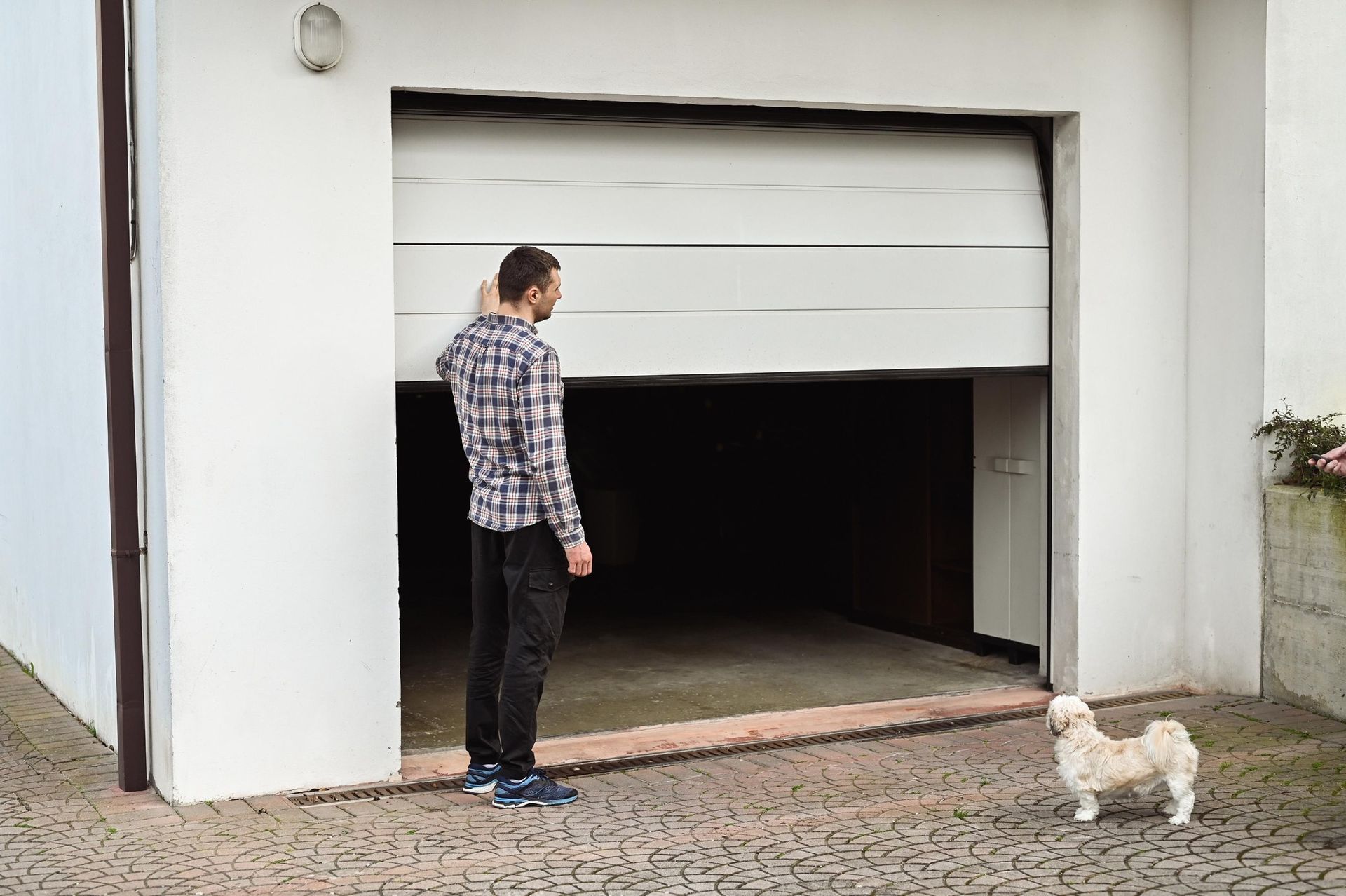 Man standing by a partially open garage door with a small dog on a cobblestone driveway