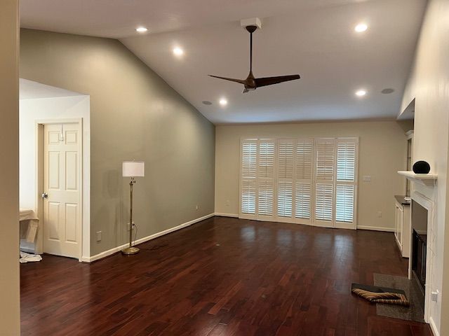 Empty living room with dark hardwood floors, vaulted ceiling, and white shutters.
