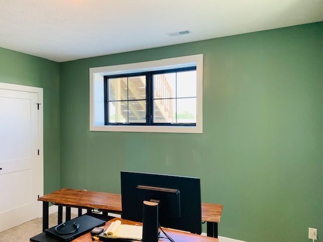 Green-walled room with a desk and window. Black-framed window with white trim, wooden desk, and white door.