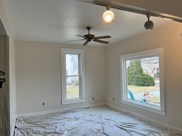 Empty room with two windows, ceiling fan, and track lighting. Pale walls and floor covered in tarp.