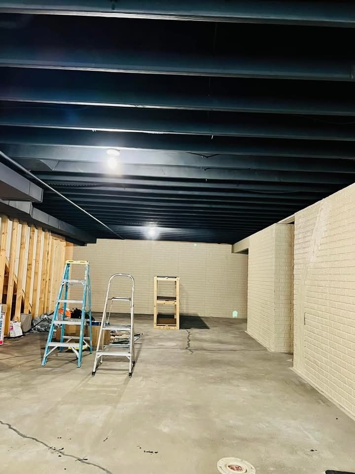 Basement interior: black ceiling beams, concrete floor, brick walls, and two ladders.