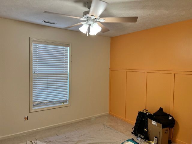 Room with an orange accent wall and tan walls. Window with blinds, ceiling fan, and carpeted floor.