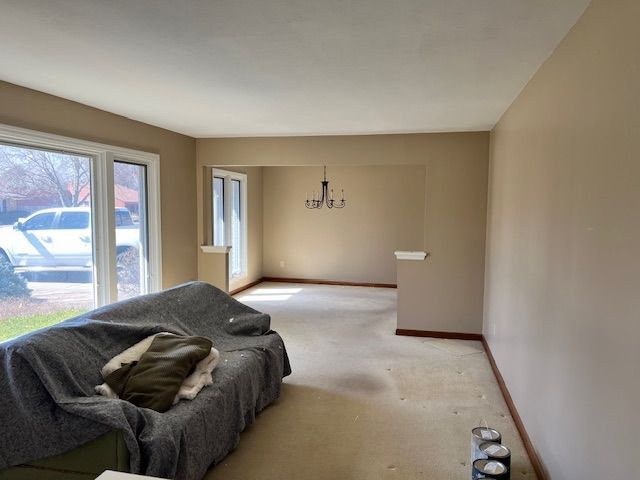 Empty living room with beige walls and carpet, covered couch, large windows, and unfinished paint cans.
