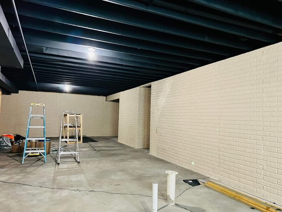 Basement renovation in progress. Exposed black ceiling beams, beige brick walls, and concrete floor. Ladders and tools present.
