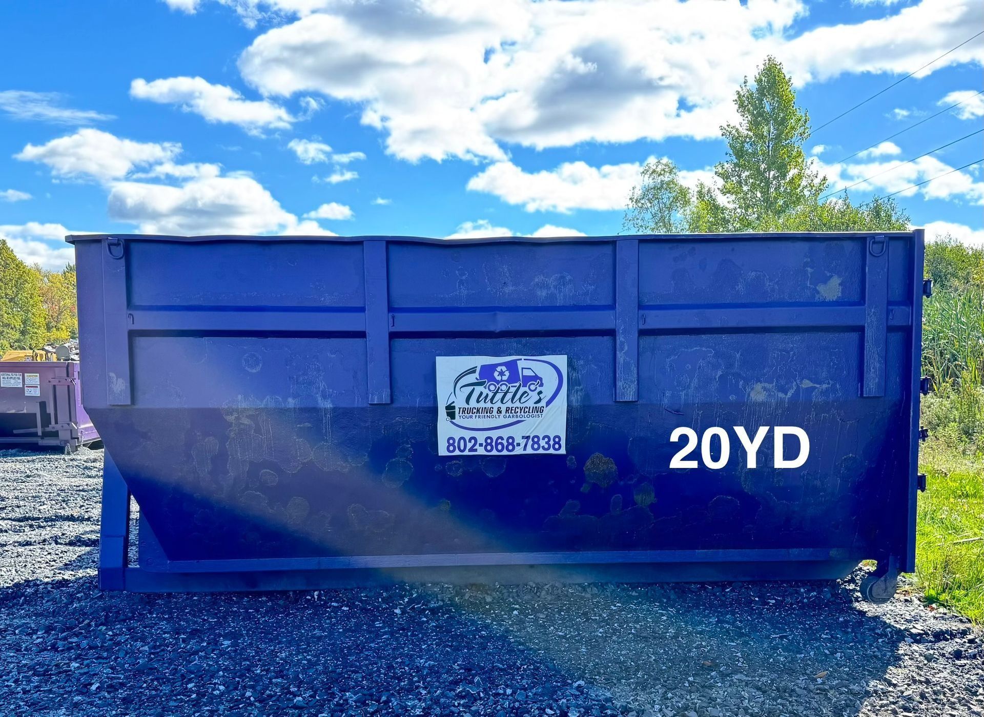 Blue 20-yard dumpster on gravel, under a blue sky with clouds.