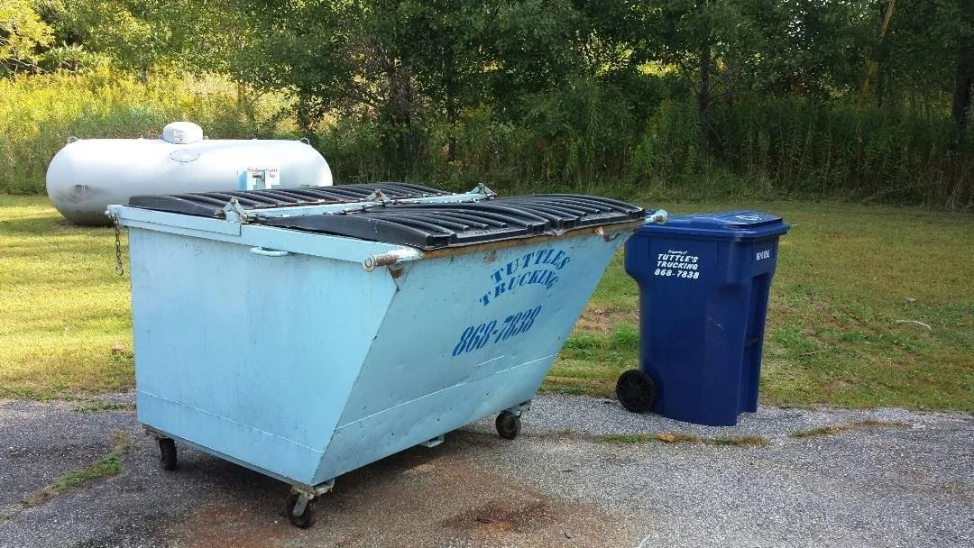 Blue dumpster with trash can next to it, and a propane tank in the background.