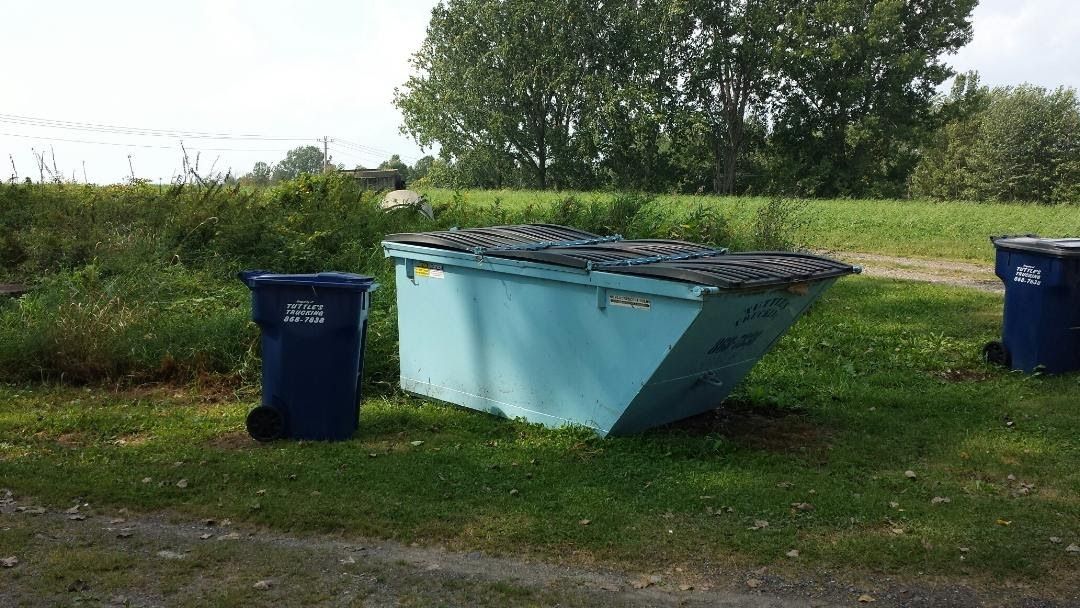 Blue dumpster and trash cans on grass near a gravel path and trees.