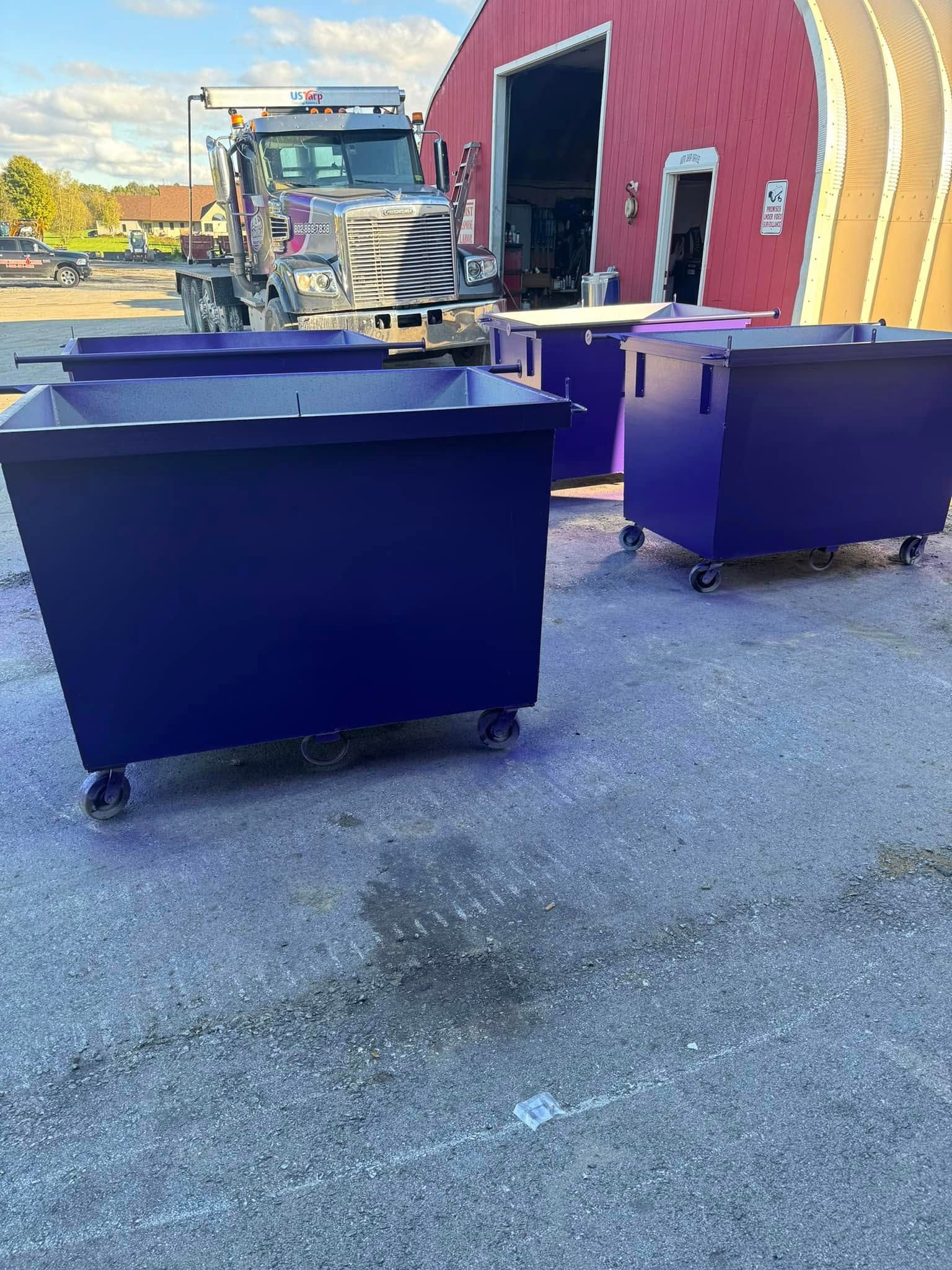 Four dark purple dumpsters on wheels in front of a red building, with a truck visible in the background.