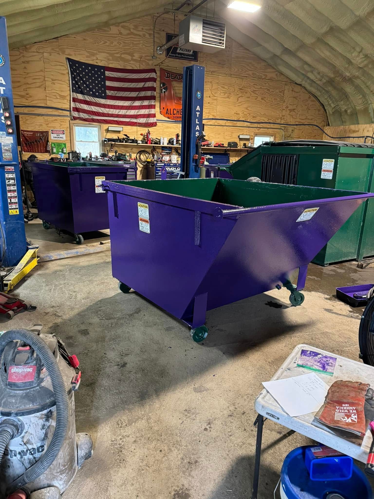 Purple and green dumpsters inside a garage with an American flag hanging on the wall.
