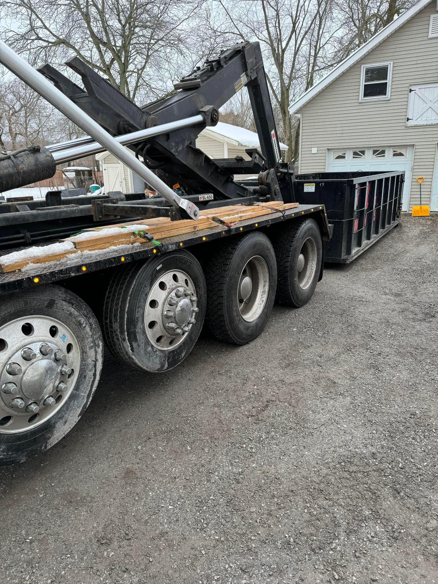 Roll-off dumpster truck on gravel driveway near a house. Black container, black truck frame, gray wheels.