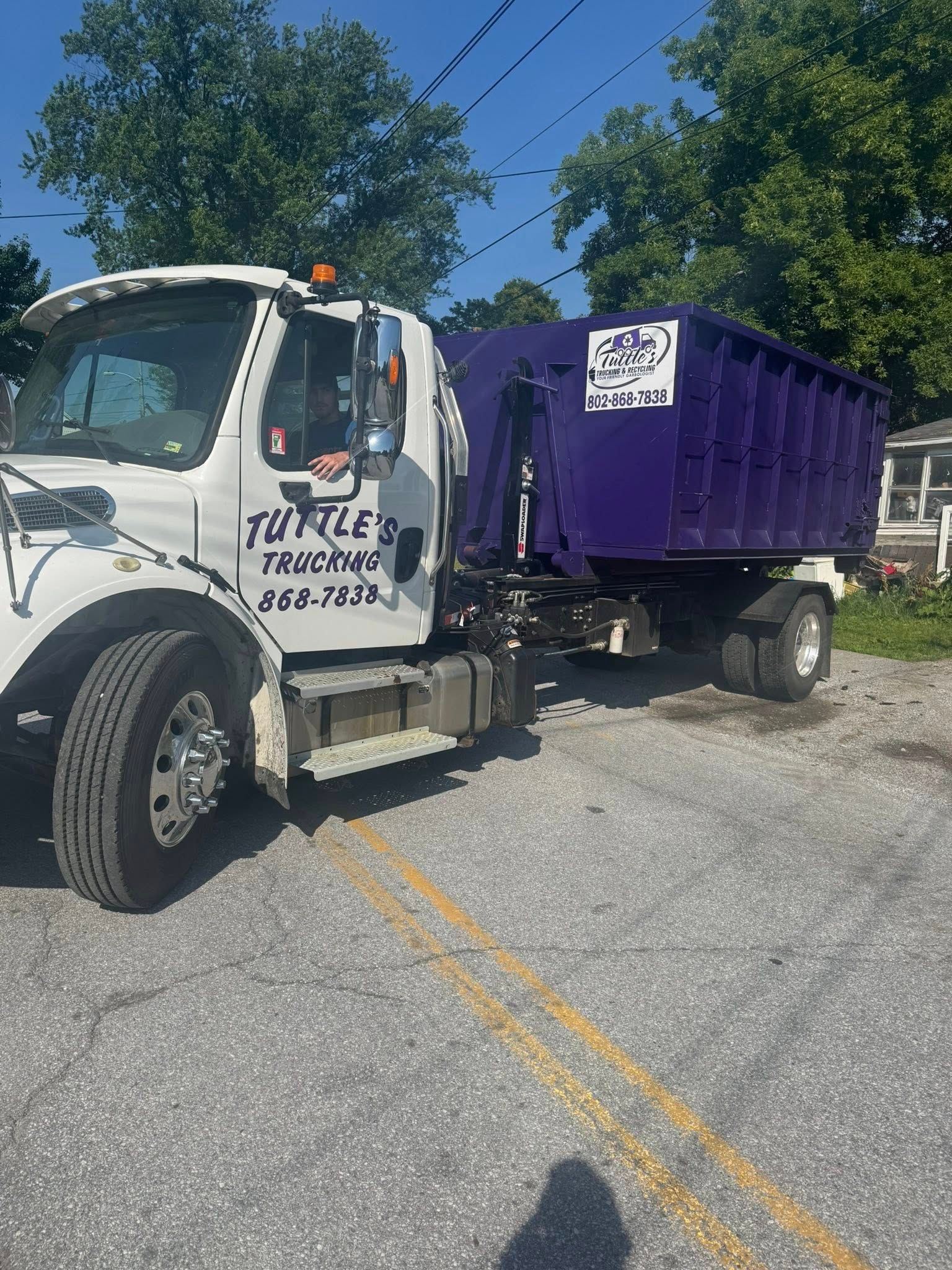 White and purple Tuttle's Trucking truck with a dumpster on a street.