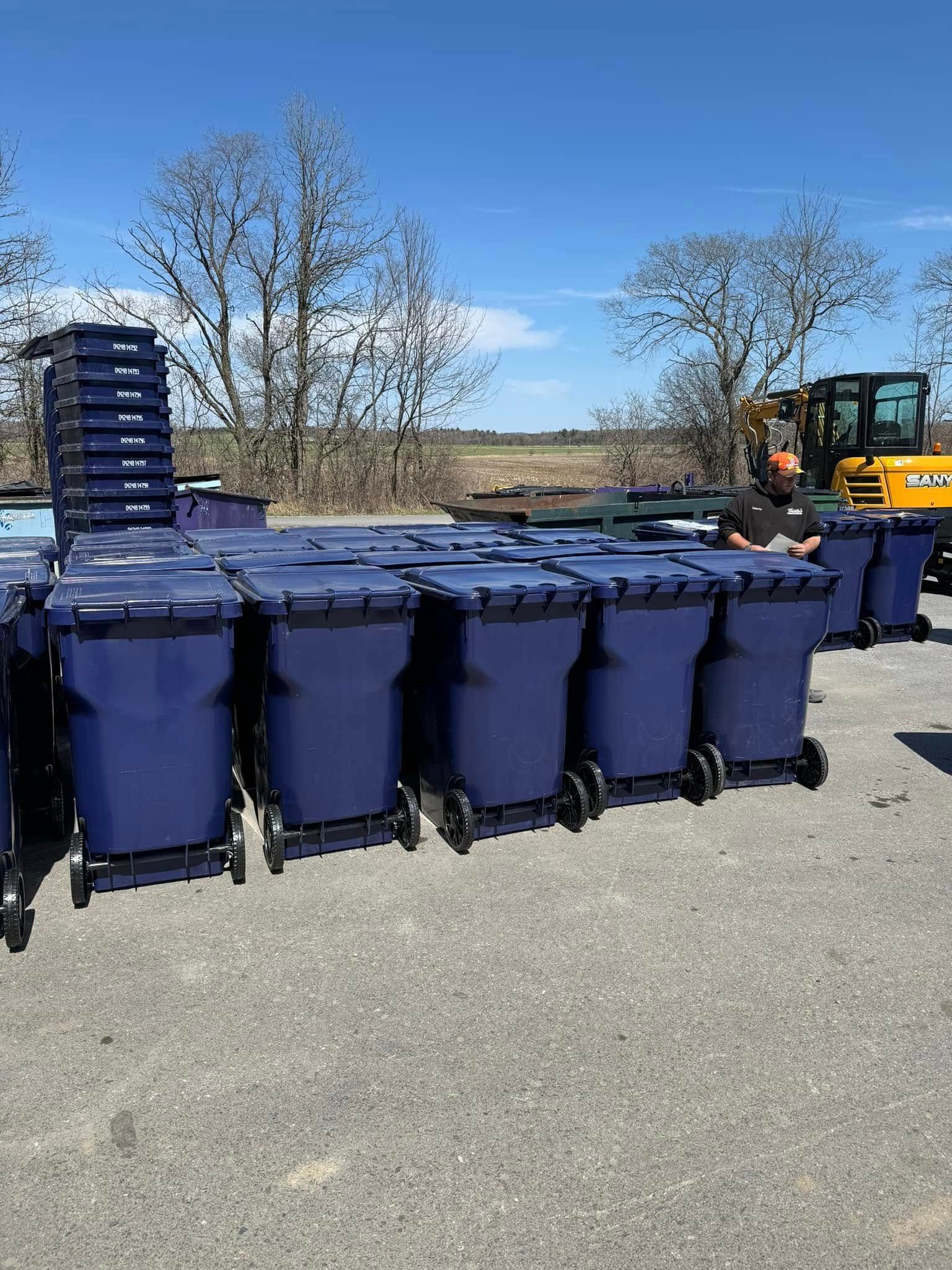 Blue trash bins with wheels, stacked and in rows, under a blue sky, near a field and construction equipment.