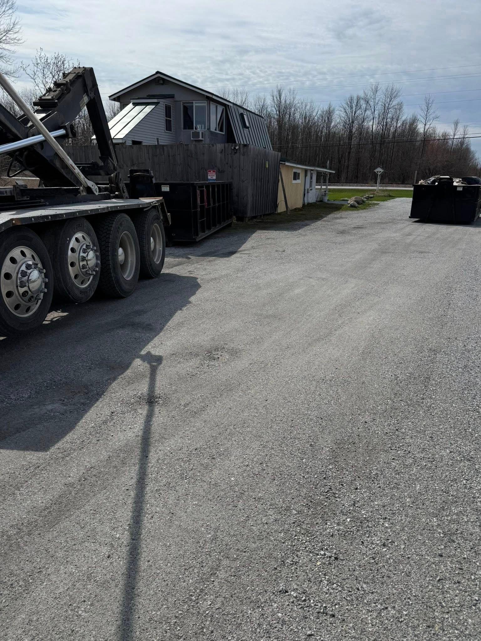 Truck with a large container next to a small, weathered building on a gravel surface.