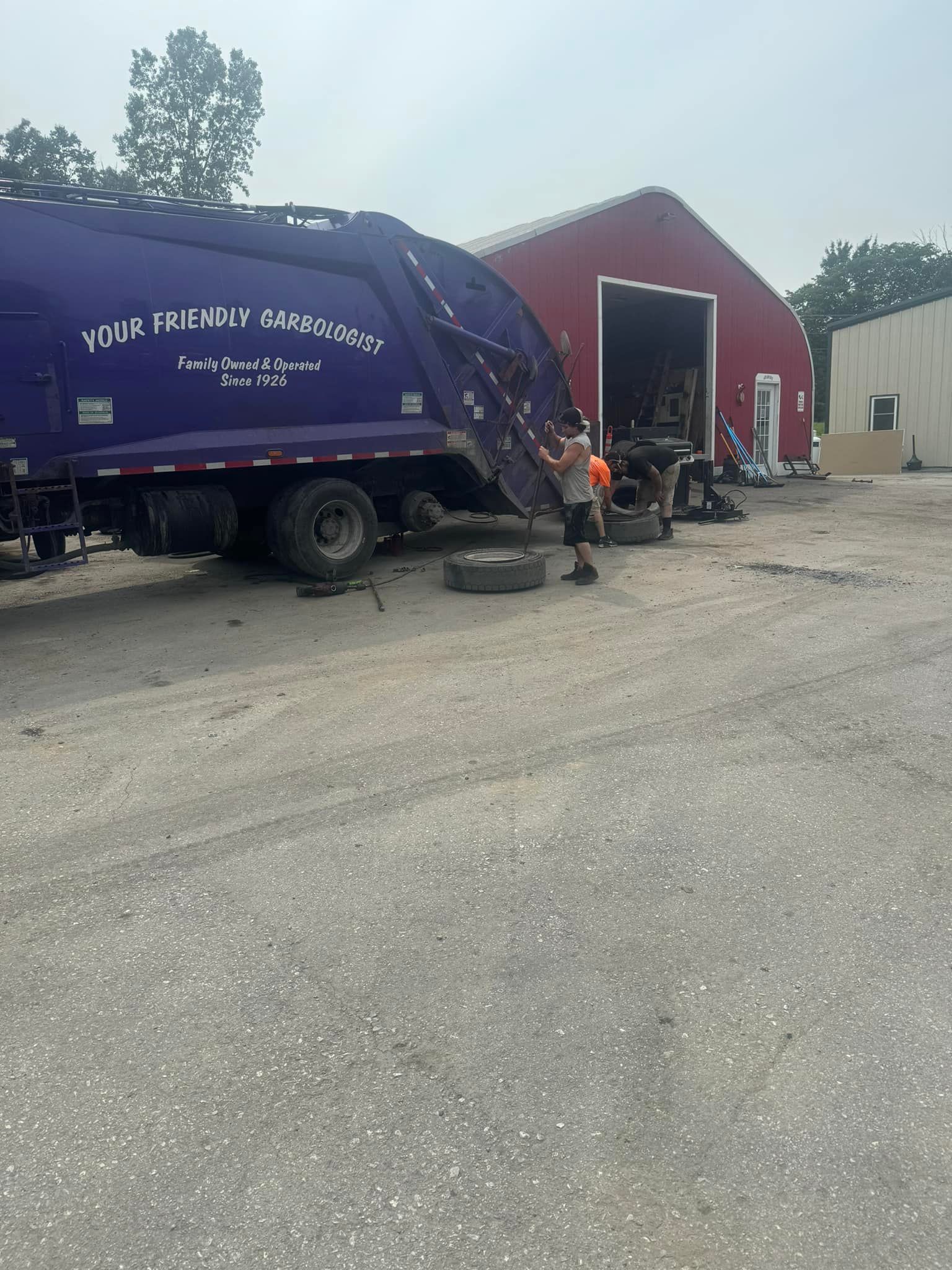 Purple garbage truck, two men changing a tire in front of a red building, outdoor setting.