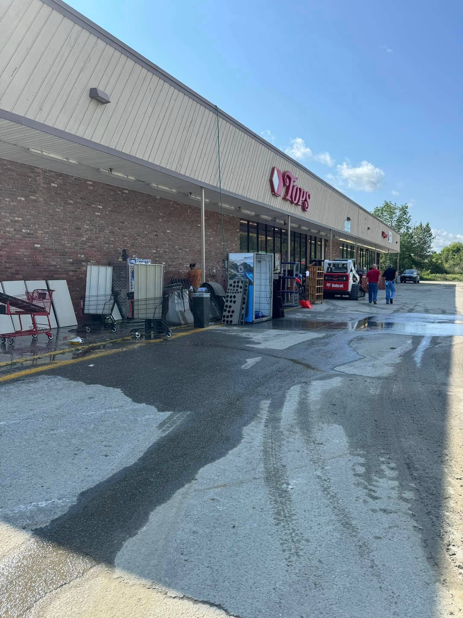 Exterior of a grocery store with water on the ground and fire trucks visible. Debris and equipment are scattered in front.